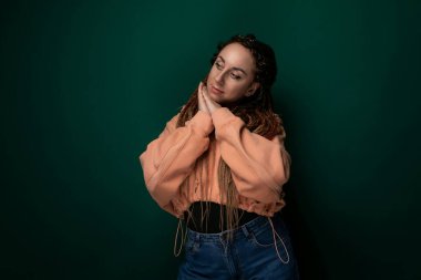 Rastalı bir kadın canlı yeşil bir duvara yaslanıyor. Kolları çapraz durduğu için rahatlamış ve dalgın görünüyor. Duvarın dokusu onun pürüzsüzlüğüyle çelişiyor.