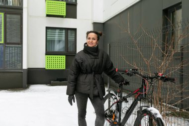 Yaşam tarzı konsepti, siyah kışlık ceketli esmer bir kadın bisikletiyle evin yanında yürüyor..