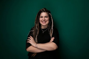 Rasta saçlı bir kadın canlı yeşil bir duvarın önünde duruyor. Sıradan giyinmiş ve kendinden emin görünüyor. Duvar onun çarpıcı görünüşü için cesur bir arka plan sağlıyor..