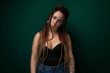 Titrek yeşil bir duvarın önünde kendinden emin bir şekilde duran rastalı bir kadın. Saçları kalın, desenli rastalarla kaplı ve güç ve bireysellik yayıyor..