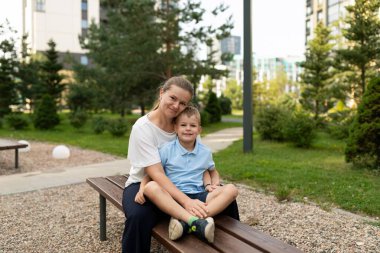 Bir anne, yeşil ve güneş ışığıyla çevrili canlı bir parktaki bankta oturup sıcak bağlarının tadını çıkarırken oğlunu kucaklıyor..