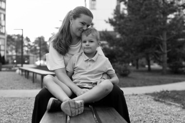 Bir anne, genç oğlunu kucağında sevgiyle tutarken onlar parkta bir bankta oturuyorlar. Atmosfer sıcaklığı ve neşeyi yansıtıyor. Açık havada huzurlu bir ortamda birbirlerine yakınlar..