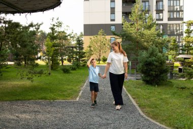 Bir anne ve oğlu ağaçlar ve yeşilliklerle çevrili canlı bir parkta el ele yürüyorlar. Neşeli etkileşimleri derin ve sevgi dolu bir ilişkiyi yansıtır..