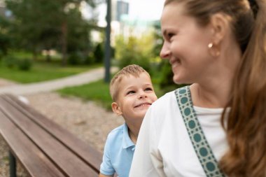 Mavi gömlekli genç bir çocuk, bir bankta oturuyor, gülümsüyor ve annesine bakıyor. Anne beyaz bir gömlek ve desenli bir kayış giyiyor ve sırtını kameraya çeviriyor..