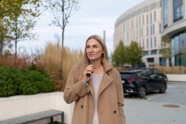 Bronz bir ceket ve gri bir kazak giyen bir kadın bir iş merkezinin önünde kulağına telefon tutarken duruyor. Ön planda ağaçlar ve çalılar var..