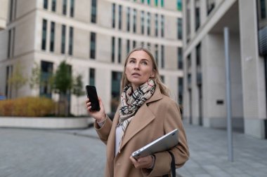 Binanın dışında bej bir ceket ve eşarp giyen bir kadın sağ elinde akıllı bir telefon ve sol elinde bir dizüstü bilgisayar tutuyor..