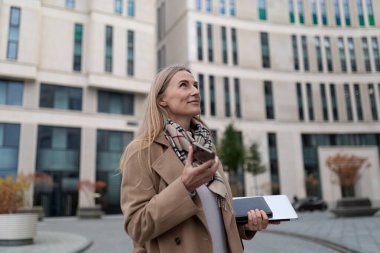 Bronz ceketli bir kadın modern bir iş merkezinin önünde duruyor, bir elinde telefon, diğer elinde dizüstü bilgisayar. Yukarı bakıyor ve düşünüyor ya da planlıyor gibi görünüyor..