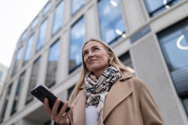 Uzun bir palto ve eşarp giyen bir kadın, geniş pencereli, uzun ve modern bir binanın önünden geçiyor. Kafasını kaldırıp elinde siyah bir akıllı telefon tutuyor..