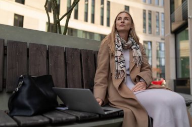 Bronz bir ceket ve atkı giyen bir kadın binanın dışındaki ahşap bir bankta oturmaktadır. Bankta siyah bir çanta onun yanında duruyor. Bir dizüstü bilgisayar kullanıyor ve çerçevenin dışında bir şeye bakıyor..