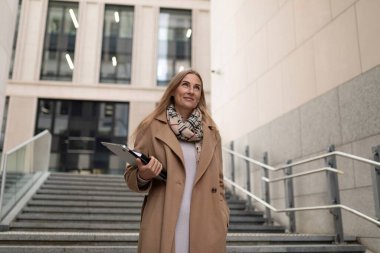 Palto giyen ve pano tutan bir iş kadını modern bir iş merkezinin merdivenlerinden yukarı çıkıyor, gülümsüyor ve iyimser bakıyor..