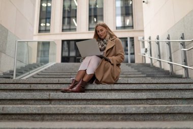 Profesyonel bir kadın, dışarıda doğal ışık altında çalışırken dikkatle dizüstü bilgisayarına odaklanmış çağdaş bir iş merkezinin merdivenlerinde oturur..