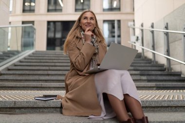 Bir iş kadını, yoğun bir iş ortamı içinde laptopuyla meşgul olan çağdaş bir ofis binasının merdivenlerinde düşünceli bir şekilde oturur..