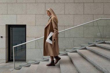 Profesyonel bir kadın iş merkezindeki taş merdivenlerden elinde dizüstü bilgisayarla güvenle iniyor ve zarif bir şekilde palto giyiyor..