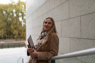 Uzun sarı saçlı bir kadın taş bir duvarın karşısında duruyor. Kahverengi bir ceket, desenli bir eşarp giyiyor ve elinde kapalı bir dizüstü bilgisayar tutuyor. Gülümsüyor ve kameraya bakıyor..