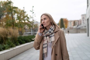 Bir kadın iş merkezinin dışında kaldırımda yürüyor. Üzerinde kahverengi bir ceket ve siyah beyaz desenli bir eşarp varken telefonuyla konuşuyor..