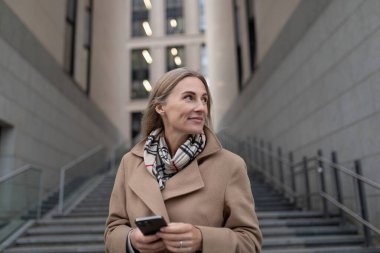 Kahverengi paltolu ve eşarplı bir kadın merdivenlerden yukarı çıkıyor. O, elinde bir telefon tutuyor ve sağına bakıyor. Arka planda, modern bir iş merkezi var..