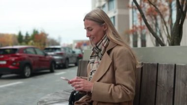 Bir kadın, şık bir ceket ve eşarp giyip arkadaşlarıyla iletişim kurmak için akıllı telefonunu kullanarak parkta rahat bir andan zevk alır. Sonbahar manzarası çevresine cazibe katıyor..