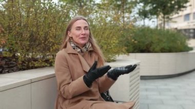 Sıcak bir palto ve eldiven giymiş bir kadın, yeşillik ve sonbahar yapraklarıyla dolu güzel manzaralı bir bankta otururken düşüncelerini ifade eder..