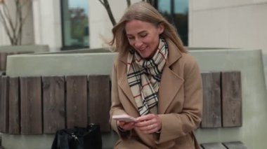 Parktaki bankta oturan bir kadın sıcak giyinmiş, güneşli bir öğleden sonranın tadını çıkarıyor. Telefonunu kontrol ediyor ve gülümsüyor. Açık bir şekilde temiz havanın tadını çıkarıyor..