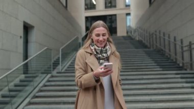 Bir kadın açık havada vakit geçirmeyi sever, akıllı telefonuyla bir dizi basamağın üzerinde dururken gülümseyerek. Modern mimari atmosfere canlılık katıyor..