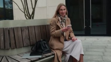 Bir kadın akıllı telefonuna bakarken rahat bir şekilde bankta oturuyor ve gülümsüyor. Şehir ortamında ağaçlar ve modern mimari, dingin bir atmosfer yaratıyor..
