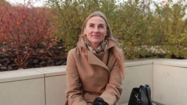 Bir kadın, sonbahar için sıcak giyimli, sakin bir parktaki bankta dinleniyor. Titrek yapraklar güzel bir zemin oluşturur. Güzel havanın tadını çıkarırken..