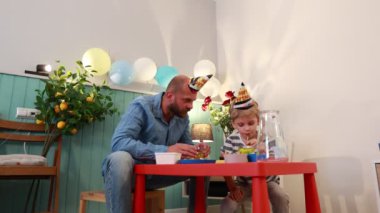 Bir baba ve oğlu küçük bir masada oturuyor, her ikisi de doğum günü şapkası takıyor. Evde balonlar ve küçük bir limon ağacı da dahil olmak üzere süslemelerle çevrili bir doğum günü kutluyorlar..