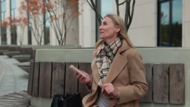 Bir kadın gülümsüyor ve bir şehir parkında telefonuna danışırken sonbahar yapraklı ağaçlarla çevrili aydınlık bir öğleden sonra.