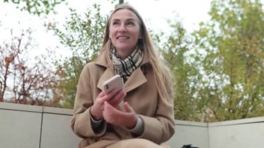 Uzun saçlı genç bir kadın, canlı bir şehir parkında renkli sonbahar yapraklarıyla çevrili telefonuyla mesajlaşırken gülümsüyor. Atmosfer neşeli ve rahat..