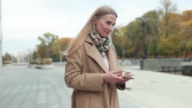 Sakin bir sonbahar öğleden sonrasında, bir kadın şehir parkında duruyor, akıllı telefonuna dalmış, etrafı renkli yapraklarla çevrili. Atmosfer sakin ve rahat..