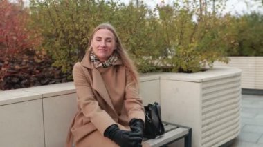 Bir kadın, sonbahar havası için sıcak giyimli, sakin bir parktaki bankta rahat bir şekilde oturur. Etrafı renkli yapraklarla çevriliyken gülümsüyor, dingin atmosferin tadını çıkarıyor..