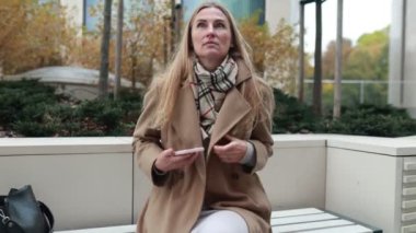 Parktaki bankta oturan bir kadın şık bir palto giymiş, yanında bir çanta dururken telefonunu tutuyor. Sonbahar yaprakları dingin bir ortam yaratır..