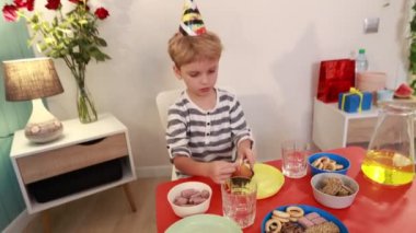 Renkli bir parti şapkası takan genç bir çocuk önünde bir kekle masada oturuyor. Kekinin üzerindeki mumu yakmak için kürdan kullanıyor..
