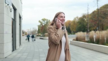 Bir kadın, sonbahar yeşillikleri ve huzurlu manzarayla çevrelenmiş bir halde şehir parkında asfalt bir yolda yürüyor, neşeli bir telefon görüşmesi yapıyor..