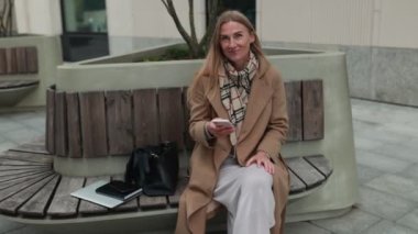 Şehirdeki bir bankta oturan genç bir kadın akıllı telefonunu kontrol ederken gülümsüyor. Şık bir ceket ve eşarp giyerek öğleden sonra atmosferinin tadını çıkarıyor..