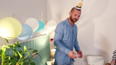 Sakallı ve kafasında doğum günü şapkası olan bir adam sarı bir balon tutuyor. Duvara bağlı bir kaç balonla birlikte bir duvarın önünde duruyor..