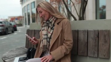 Şık bir ceket giymiş bir kadın, sonbaharda kalabalık bir şehir parkında otururken telefonunda bir mesaj okurken heyecanla tepki veriyor..
