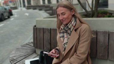 Uzun saçlı neşeli bir kadın günümüzün kentsel ortamında bankta oturuyor, bej bir ceket giyiyor ve elinde küçük bir nesneyle mutlu bir şekilde gülümsüyor..