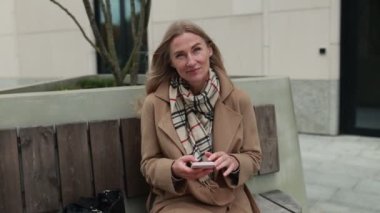 Şık bir palto ve eşarp giymiş bir kadın, şehir parkında güzel bir sonbahar havasının tadını çıkarırken telefonuna bakarak bankta dinleniyor..