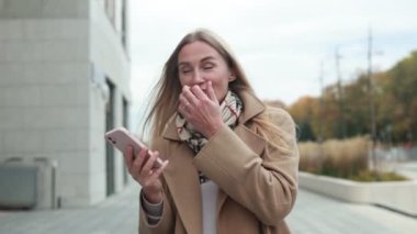 Neşeli bir kadın, sonbaharda şehir merkezinde dolaşırken akıllı telefonundan zevk alır. Saçlarını düzeltip kameraya gülümserken pozitiflik saçıyor..