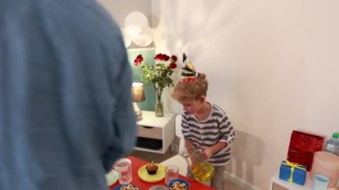 Doğum günü şapkası takan genç bir çocuk balon ve abur cuburla süslenmiş bir masaya meyve suyu döker. Babasıyla evde kutlama yapıyor..