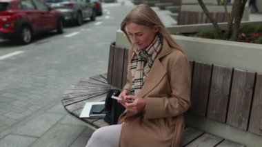 Şehirde ahşap bir bankta oturan zarif bir kadın telefonuna odaklanmış. Uzun binalar ve park halindeki arabalar sonbaharda huzurlu bir şehir arkaplanı oluşturuyor.