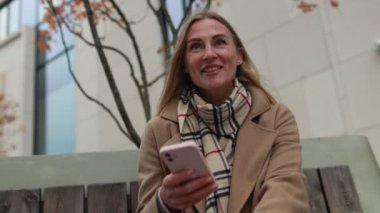 Bir kadın, şehir parkındaki bir bankta rahat bir şekilde oturup, akıllı telefonuyla sonbahar atmosferinin tadını çıkarıyor ve etrafındaki manzaranın tadını çıkarıyor..