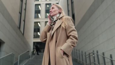 Moda sahibi bir kadın, modern mimariyle çevriliyken bir merdivende telefon görüşmesi yapıyor. Sıkı giyinmiş, açık günün tadını çıkarıyor..