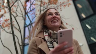 Bir kadın akıllı telefonuyla meşgul, gülerek kentte sonbaharın güzelliğinin tadını çıkarıyor. Etrafını saran renkli yapraklar neşe dolu atmosfere katıyor..