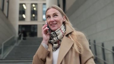 Şık bir ceket ve eşarp giyen bir kadın, çağdaş bir şehir ortamında beton merdivenlerden inerken telefonuyla neşeli bir şekilde konuşur..