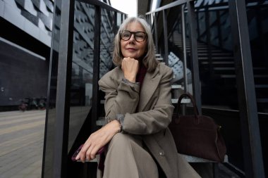 Yaşlı bir kadın zarif bir şekilde merdivenlerde oturur, modern bir binanın dışında beklerken çenesini elinin üzerine koyar. Şehir çevresi onun dengeli tavrını geliştiriyor..