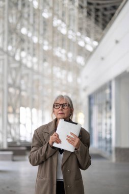Yaşlı bir iş kadını, günümüzün ofis ortamında elinde bir pano tutarak, hareketli bir atmosferde sonraki adımlarını düşünerek düşünceli bir şekilde duruyor..