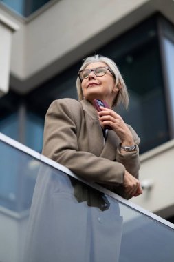 Yaşlı bir iş kadını zarif bir şekilde balkon korkuluklarına yaslanıyor, şehrin ufuk çizgisine dikkatlice bakıyor. Öğleden sonra güneşi üzerinde sıcak bir parıltı yayar, düşünceli ifadesini güçlendirir..