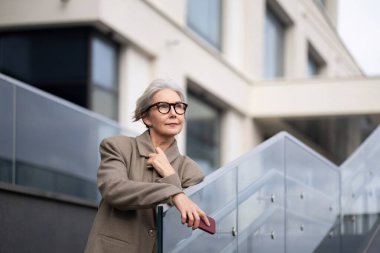 Moda gözlüklü yaşlı bir kadın, arka planda açık gökyüzünün altında modern bir bina bulunan çağdaş bir merdivende düşünceli bir şekilde poz veriyor..